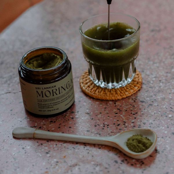 Moringa powder being whisked into a glass of water with an electric frother, creating a vibrant green drink, with the moringa jar and a ceramic spoon in view.