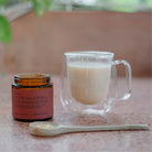 A prepared creamy ashwagandha latte in a clear glass mug, placed next to an open jar of ashwagandha powder and a ceramic spoon with powder.