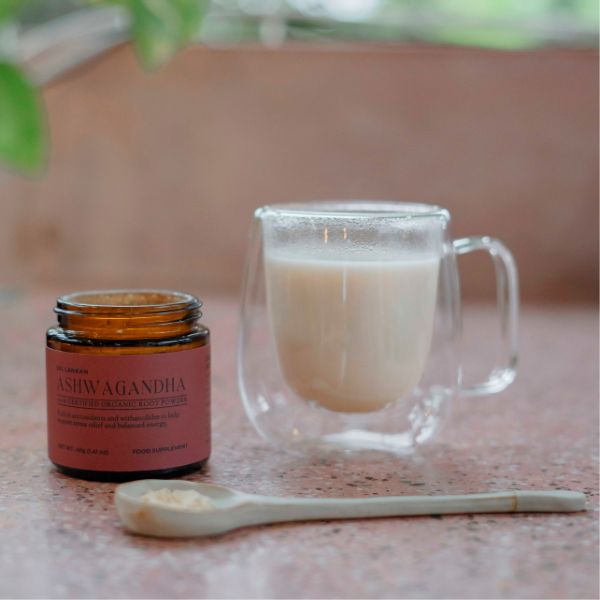 A prepared creamy ashwagandha latte in a clear glass mug, placed next to an open jar of ashwagandha powder and a ceramic spoon with powder.