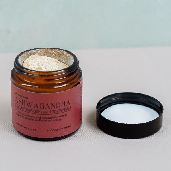 An open glass jar of light beige ashwagandha root powder, with the black lid placed beside it on a neutral surface."