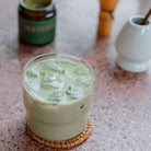 A glass of iced matcha latte with creamy green color, topped with ice cubes, sitting on a woven coaster with a blurred jar of matcha in the background.