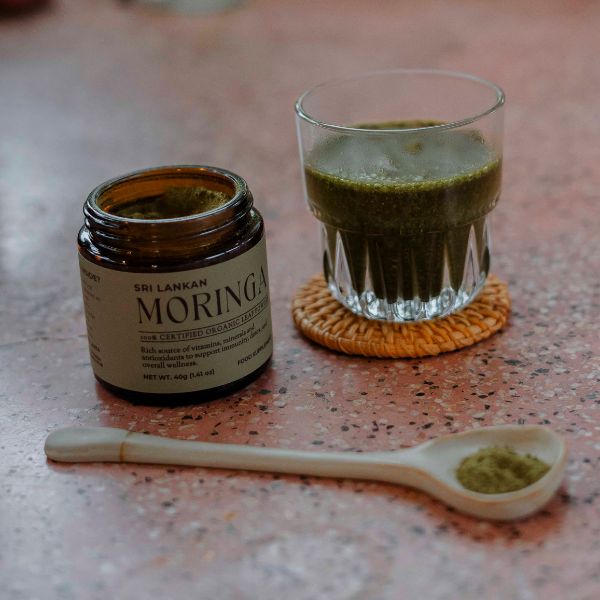 A freshly prepared glass of moringa drink, vibrant green in color, placed on a woven coaster next to an open jar of moringa and a ceramic spoon with powder."