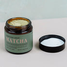 An open jar of Sri Lankan ceremonial-grade matcha powder with a black lid placed beside, showing vibrant green matcha powder inside.