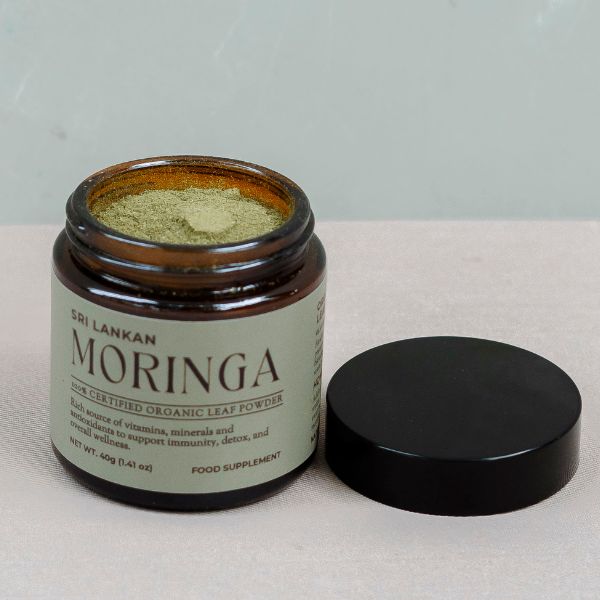 An open glass jar of fine, vibrant green moringa powder, with the black lid placed beside it on a neutral surface.