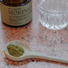 A ceramic spoon holding a small amount of fine, green moringa powder, with the moringa jar and a glass in the background.