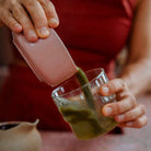 A person carefully pouring freshly whisked ceremonial-grade matcha from a pink bowl into a clear glass for a vibrant green drink.