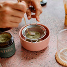 Hands sifting ceremonial-grade matcha powder through a fine mesh sieve into a pink bowl, highlighting the delicate, finely ground texture of the matcha.