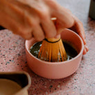 A hand using a bamboo matcha whisk to mix ceremonial-grade matcha with water in a pink bowl, forming a smooth, frothy texture.